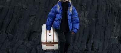 woman walking on a cliff holding the Manor large hardshell luggage in the color white with brown trim.