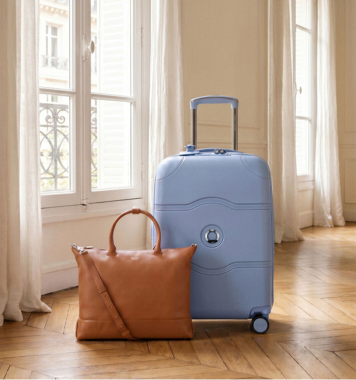 Light blue Delsey carry-on suitcase and a tan leather tote on a wooden floor by tall windows